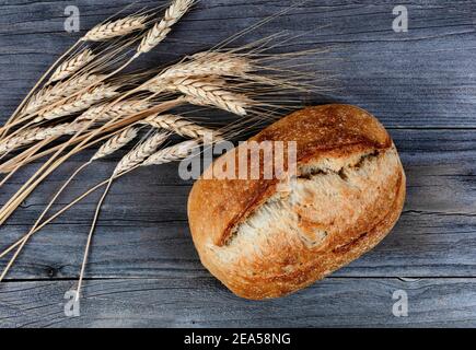 Close up view of homemade whole sourdough loaf of bread with dried wheat stalks on weathered wooden planks