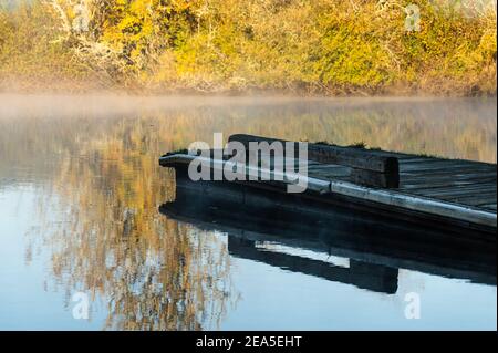 Boat dock in river surrounded by bright orange trees with fog on water Stock Photo