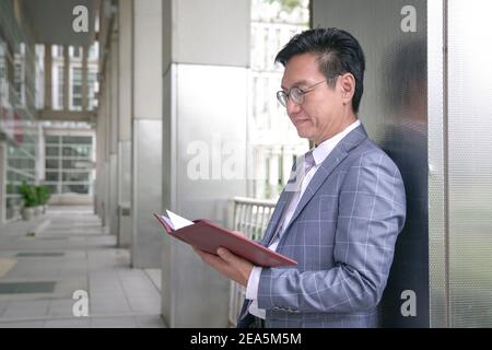 Asian businessman in jacket leaning against building pillar and looking at his diary Stock Photo