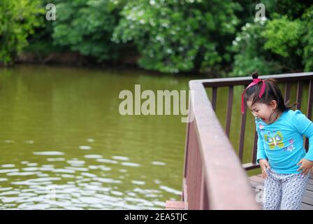 A cute young Asian girl, standing on a bridge by a pond, looking down at the water and feeling excited after seeing a turtle. Jumping up and down with Stock Photo