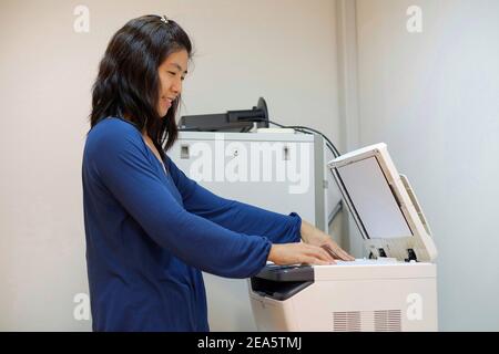 A middle aged Asian woman, working as a secretary is copying documents using a multifunction laser printer at her office. Stock Photo