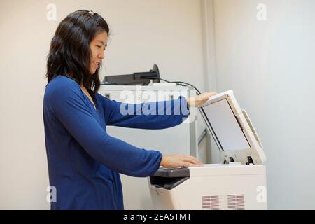 A middle aged Asian woman, working as a secretary is copying documents using a multifunction laser printer at her office. Stock Photo