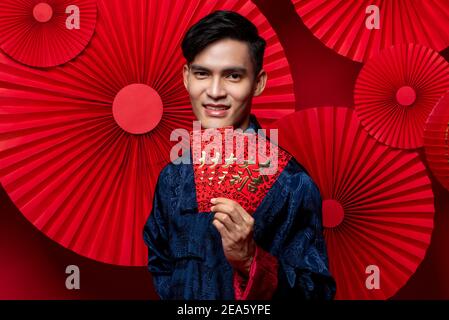 Young Asian man in traditional costume holding red envelopes or Ang Pao as a gift for Chinese new year in red oriental style background, foreign text Stock Photo