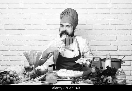 Chef kneads dough. Cook with excited face in burgundy uniform sits by table with vegetables and kitchenware. Cooking process concept. Man with beard licks dough off fingers on white brick background Stock Photo