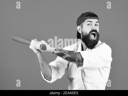 Hooligan gets ready to fight. Karate man with angry face holds green baseball bat. Violence and bullying concept. Man with beard in white kimono and green cap on blue background. Stock Photo
