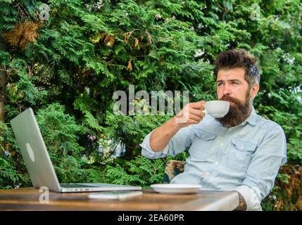 Pleasant moment. Take moment to enjoy life. Man bearded hipster make pause for drink coffee and relax while sit with laptop. Guy drinks coffee relaxing terrace branches background. Aroma cappuccino. Stock Photo