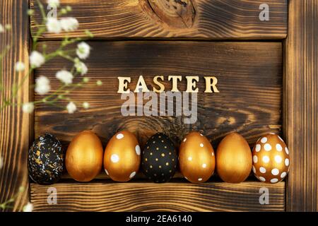 Easter festive background. Top view of easter eggs colored with golden paint and inscription in English Easter. Wooden letters on dark wood background Stock Photo