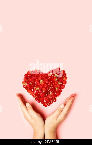 Beautiful fruit heart made of berries with female hands on a pink background with copy space. View from above. Vertical photo Stock Photo