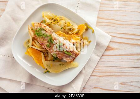 Cod fish fillet on fennel vegetable with orange slices and fennel green garnish on a white plate and napkin, light wooden table with copy space, high Stock Photo
