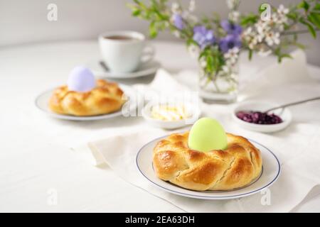 Easter breakfast with plaited yeast bread nests and colored eggs, coffee, butter, jam and flowers on a white table, copy space, selected focus, narrow Stock Photo