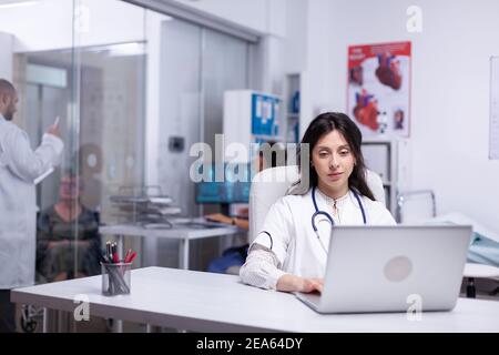 Professional doctor in white coat working on laptop in modern hospital office room, therapist typing on computer consult patient online, makes research, analysing results information from internet Stock Photo