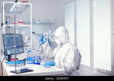 Researcher spikes samples using an automatic pipette wearing protecton suit against coronavirus. Chemist in modern laboratory doing research using dispenser during global epidemic with covid-19. Stock Photo
