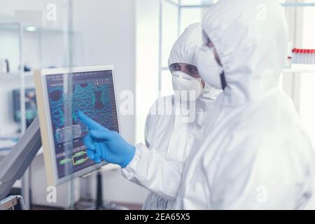 Chemist in ppe suit explainig to doctor vaccine development in modern equipped laboratory pointing on computer desktop. Doctors examining virus evolution using high tech researching diagnosis against covid19 Stock Photo