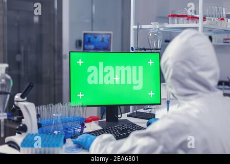 Over shoulder shot of doctor working on coroanvirus vaccine using computer with green screen dressed in ppe. Team of microbiologists doing vaccine research writing on device with chroma key, isolated, mockup display. Stock Photo