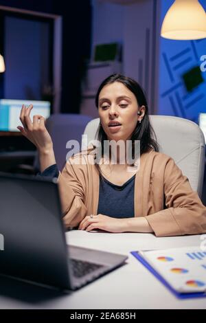 Businesswoman using earbuds in the course of online call doing overtime. Woman working on finance during a video conference with coworkers at night hours in the office. Stock Photo