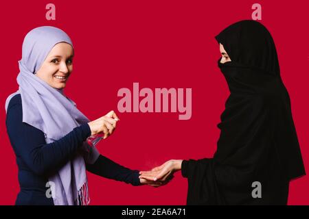 Two Muslim female friends with scarves covered their heads, treat their hands with antiseptic spray against the spread of coronavirus. Red background. Stock Photo