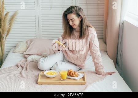 A beautiful woman sits on a bed with a healthy breakfast and communicates on the phone in messages. Stock Photo