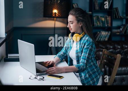 A young woman sits in a cozy interior at a table by the window and types on a laptop, working remotely from home. Distance learning, the use of modern Stock Photo