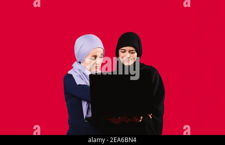 Work online. Concentrated Muslim female friends are working on a laptop on a red background with blank space. Stock Photo