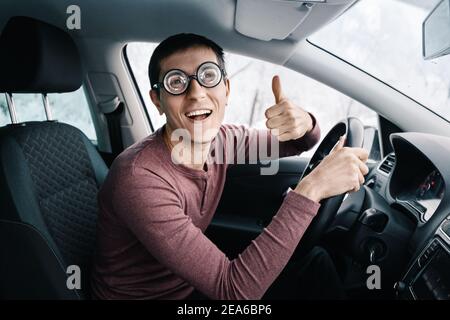 Funny and ridiculous idiotic nerd driver in big eyeglasses holding the steering wheel and smiling to the camera. Concept of a newbie driver distracted Stock Photo