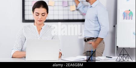businesswoman using laptop near coworker putting sticky notes on board on blurred background, banner Stock Photo