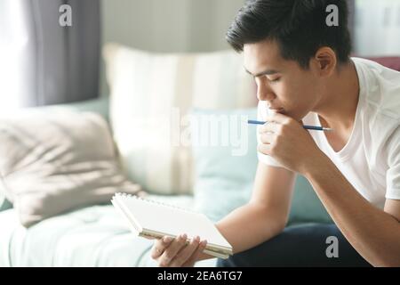 thoughtful asian young teenage student man teenager thinking holding notebook on sofa at home Stock Photo
