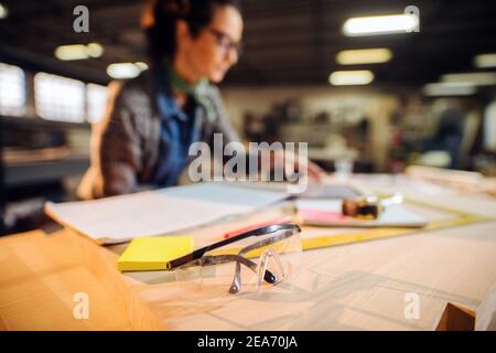 Female doing calculations in carpenter's workshop. Selective focus on protective eyewear. Stock Photo