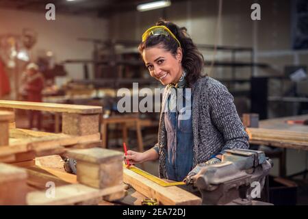 Middle aged professional female carpenter looking at the camera while taking measures of raw wood in her workshop. Stock Photo