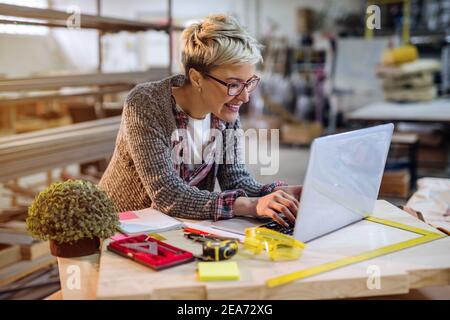 Female carpenter using laptop for calculations while standing in the workshop. Stock Photo
