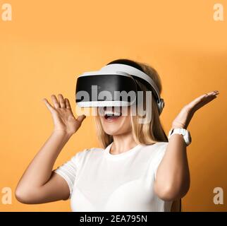 Portrait of happy screaming blonde woman in white t-shirt using VR glasses exploring augmented reality holding hands up Stock Photo