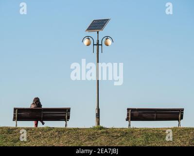 Lonely woman sitting on a bench next to a light pole. Two benches with a solar panel lamp post in the middle Stock Photo