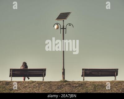 Lonely woman sitting on a bench next to a light pole. Two benches with a solar panel lamp post in the middle Stock Photo