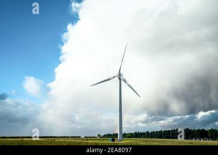 Single lone electric generating wind turbine windmill with dramatic big storm cloud and rainbow and nature park forest woodland behind on bright day Stock Photo