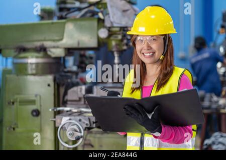 Portrait of Asian woman sales engineer checking the job list in paper over the photo blurred of lathe and milling machine background in metal factory, Stock Photo
