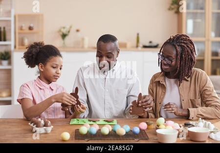 Portrait of loving African-American family making chocolate Easter bunnies while sitting at wooden table in cozy home interior and enjoying holiday pr Stock Photo