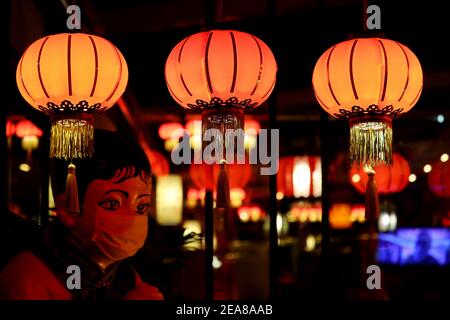 Red Chinese paper lanterns glow in a row on a night street on restaurant entrance and statue in medical mask background. New Year celebration in China Stock Photo
