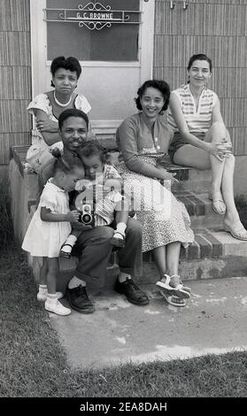 1960s, Chicago, USA, a family siiting on steps of their property, with the two youngest daughters taking an interest in their father's film camera, a Ciroflex, a twin-lens reflex camera (TLR). The Ciro Camera co started making cameras in Detroit, in 1941 and then in Delaware, Ohio, from 1947. In 1951 Graflex acquired the rights to make the camera and produced the Ciro flex twin-lens reflex.  Six models were made, the one here, the Model E, was known as the 'working man's Rolleiflex', as it was affordable for the man in the street. Stock Photo