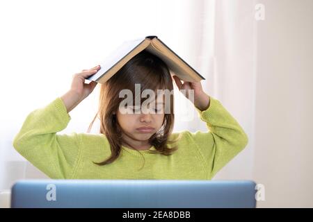 Sad elementary schooler girl studying at home with a book on her head because she is tired and angry - Little girl studying online and using internet Stock Photo