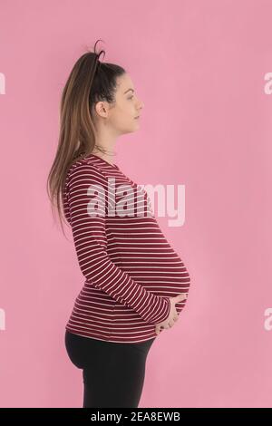 Focused looking pregnant woman holding her belly on a pink background. Side view. Pregnancy concept. Stock Photo