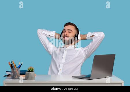 Taking break from work. Smiling Caucasian man listening to music in headphones at his desk on blue studio background Stock Photo