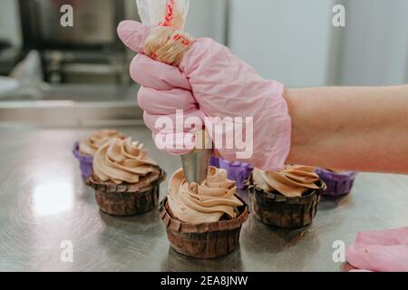 Close up of female confectioner wearing pink gloves making cupcakes using pastry bag with cream in the confectionery studio Stock Photo