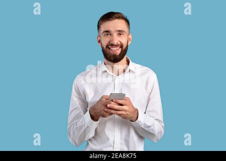 Cheerful millennial guy with smartphone browsing web, playing game, shopping online on blue studio background Stock Photo