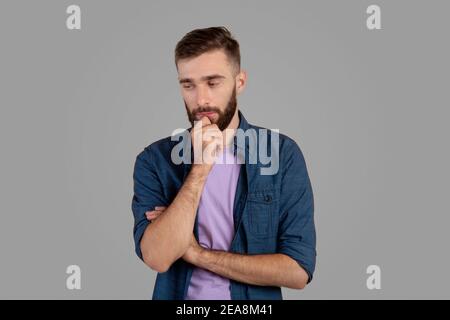 Dreamy young guy thinking over problem, touching his chin on grey studio background Stock Photo
