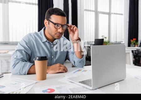 Indian businessman looking at laptop near coffee to go and papers on blurred foreground Stock Photo