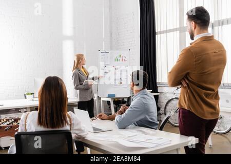 Businesswoman with digital tablet pointing at graphs on flipchart near multiethnic colleagues on blurred foreground Stock Photo