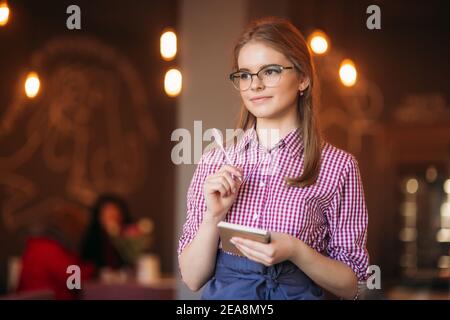 Portrait of a beautiful friendly waitress in glasses with notepapers and pen ready to take your order Stock Photo