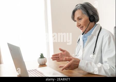 Senior old female doctor with stethoscope, wearing lab coat and headset, sitting in the office and discussing medical case with colleagues online Stock Photo