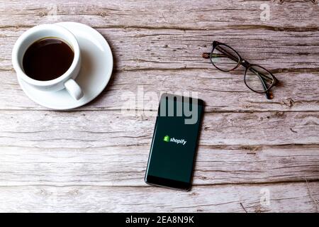 A mobile phone or cell phone laid on a wooden table with the shopify app open next to a coffee and glasses Stock Photo