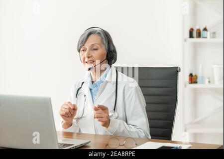 Portrait of a senior doctor with stethoscope in a lab coat sitting at the desk, wearing headset and looking at the laptop,giving consultation to a Stock Photo