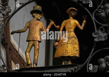 Golden couple.  Gilded centrepiece of an old metal shop sign on an early-18th century timber-framed house in Michelstadt, Hesse, Germany.  The gentleman wears a top hat, has a walking stick hooked over his arm and holds a large tobacco pipe.  The lady wears a summer dress and hat, carries a small handbag in one hand and holds up a bunch of flowers in the other. Stock Photo